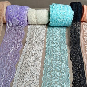 May include: A collection of eight rolls of decorative lace trim in various colors including peach, lavender, cream, turquoise, black, and white. Each roll features a distinct floral or scalloped pattern with intricate openwork detailing.