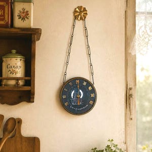 May include: A vintage-style wall thermometer with a dark blue face and gold-colored accents. The thermometer is suspended by a chain from a decorative gold flower. The face displays temperature markings and an illustration.