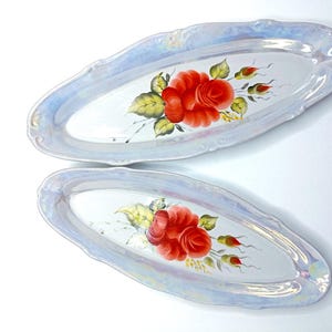 May include: Two oval serving platters with a pearlescent finish and scalloped edges. Each platter features a hand-painted red rose with green leaves and buds. The background is white.