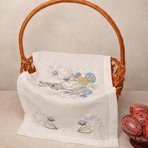May include: A wicker basket with a handle contains a white linen towel with an embroidered rabbit and floral design in blue and yellow. Decorative Easter eggs in red and black are placed nearby.
