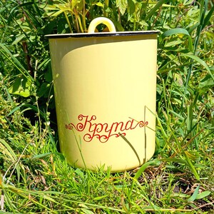 Könnte beinhalten: Ein gelber Emaillebehälter mit Deckel und Griff, der in grünem Gras steht. Der Behälter hat einen schwarzen Rand und ein rotes Schriftwort an der Seite. Der Behälter ist zylindrisch geformt.