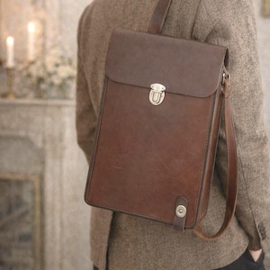 May include: A rectangular brown leather backpack featuring a fold-over flap with a metallic push-lock closure. The bag includes a vertical strap detail with a circular metal snap button at the bottom and an adjustable shoulder strap.