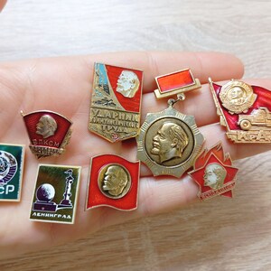 Könnte beinhalten: Eine Sammlung von acht sowjetischen Anstecknadeln aus Metall mit verschiedenen Motiven, darunter Porträts von Wladimir Lenin, das Hammer-und-Sichel-Emblem, rote Flaggen und kyrillische Schriftzüge wie CCCP, Leningrad und KRAZ in Gold und Emaille.