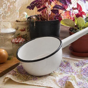 May include: A white enamel saucepan with a black rim and a long handle. The round cooking vessel features a smooth, glossy finish and a simple, functional design. The interior and exterior are white, while the edge is outlined in black.