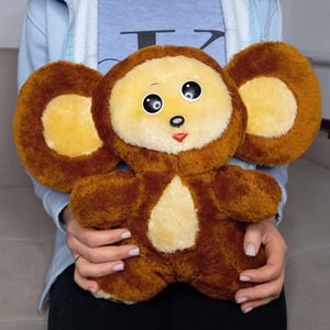 Op de afbeelding: Een pluche speelgoed in de vorm van een stripfiguur, met een bruin lichaam, grote ronde oren en een lichtgeel gezicht. Het speelgoed heeft zwarte ogen en een kleine rode mond. Het speelgoed wordt door een persoon vastgehouden.