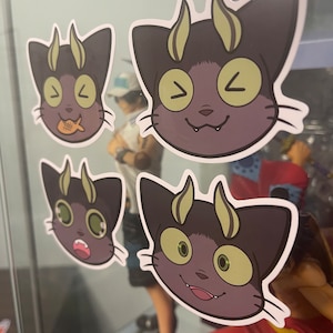 May include: Four stickers featuring a cartoon cat with horns and different facial expressions. The cat is black with green eyes and yellow horns. The stickers are on a glass surface.