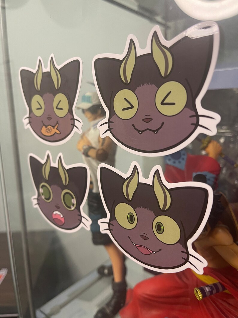 May include: Four stickers featuring a cartoon cat with horns and different facial expressions. The cat is black with green eyes and yellow horns. The stickers are on a glass surface.