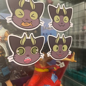 May include: Four stickers featuring a cartoon cat with horns and different facial expressions. The cat is purple with yellow horns and green eyes. The stickers are on a glass surface.