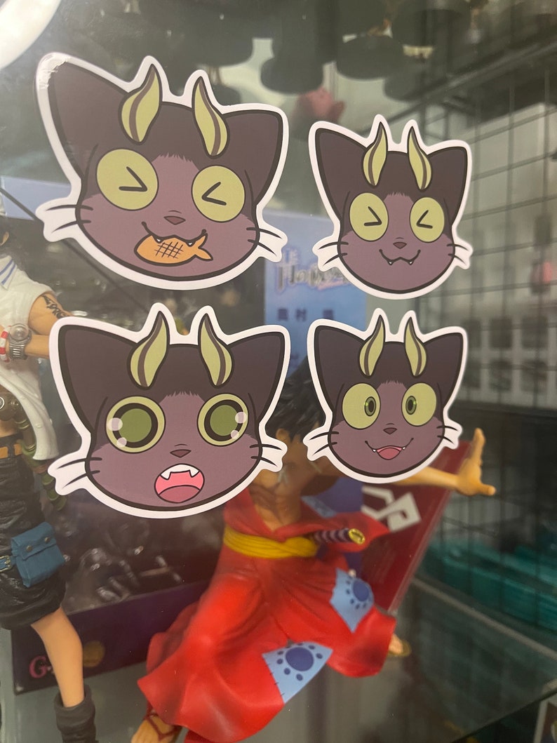 May include: Four stickers featuring a cartoon cat with horns and different facial expressions. The cat is purple with yellow horns and green eyes. The stickers are on a glass surface.