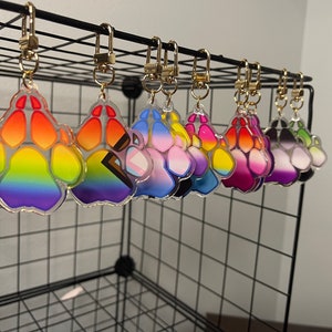 May include: A set of eight colorful paw print keychains, each with a different gradient design. The keychains are made of clear acrylic and feature a gold metal ring.