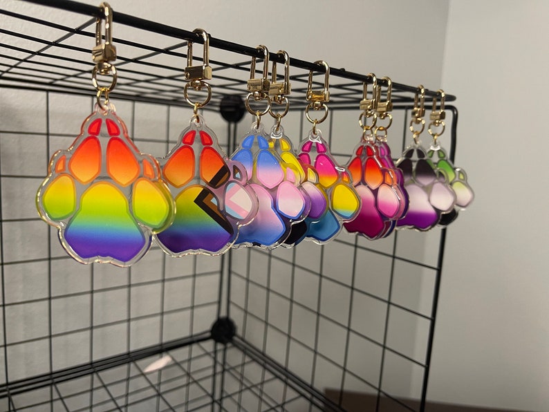 May include: A set of eight colourful paw print keyrings, each with a different gradient design. The keyrings are made of clear acrylic and feature a gold metal ring.
