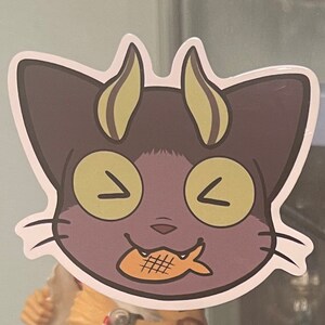 May include: A purple cat with yellow eyes and horns is smiling and holding a fish-shaped snack in its mouth.