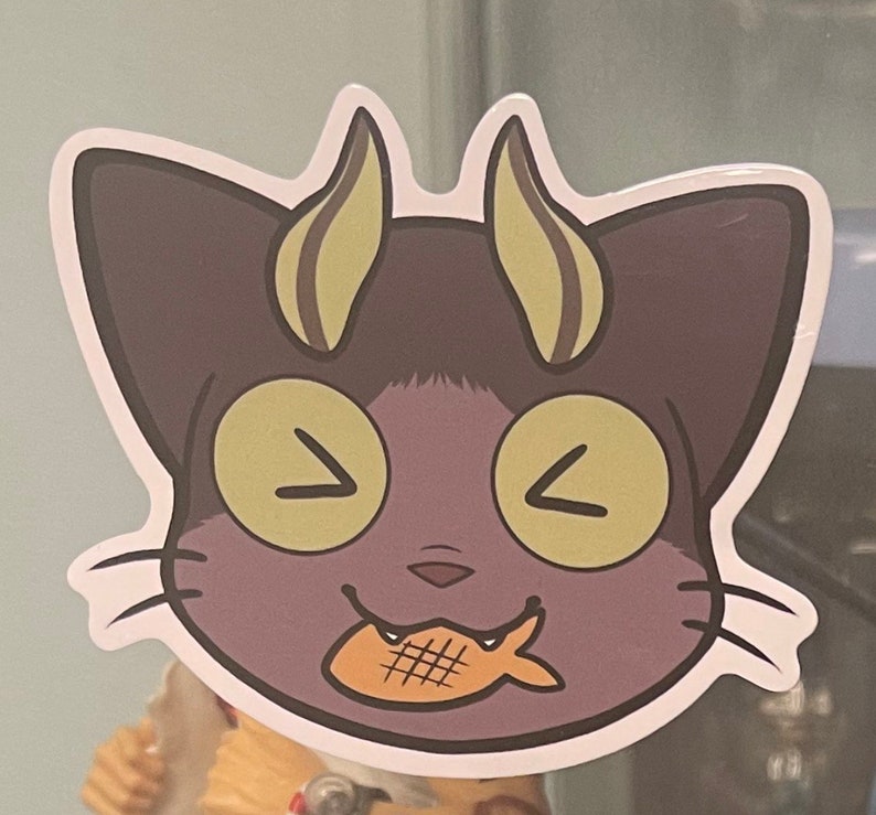 May include: A purple cat with yellow eyes and horns is smiling and holding a fish-shaped snack in its mouth.