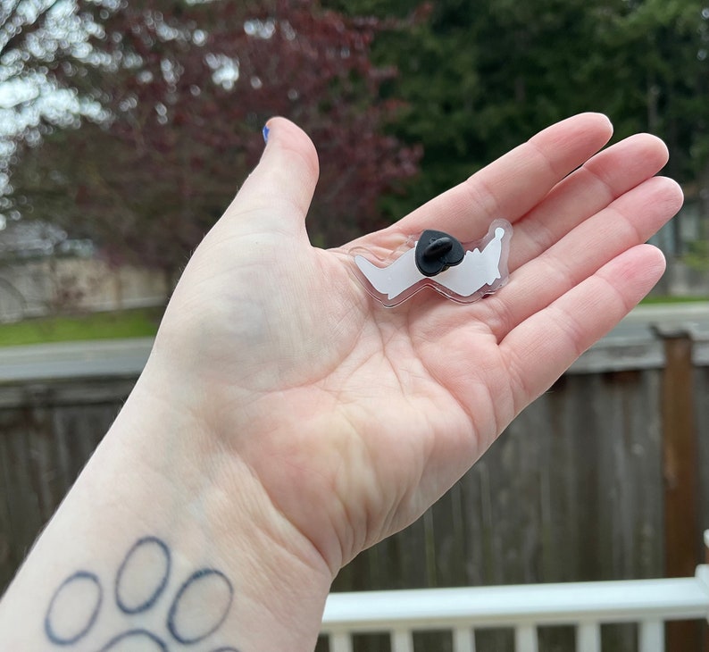 May include: A clear plastic pin with a white outline of a dog paw print. The pin has a black metal backing with a safety pin closure.