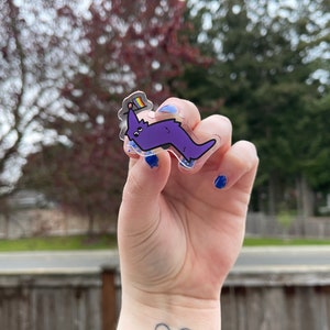 May include: A clear acrylic sticker with a purple cartoon worm wearing a rainbow flag hat. The sticker is being held by a hand with blue nail polish.