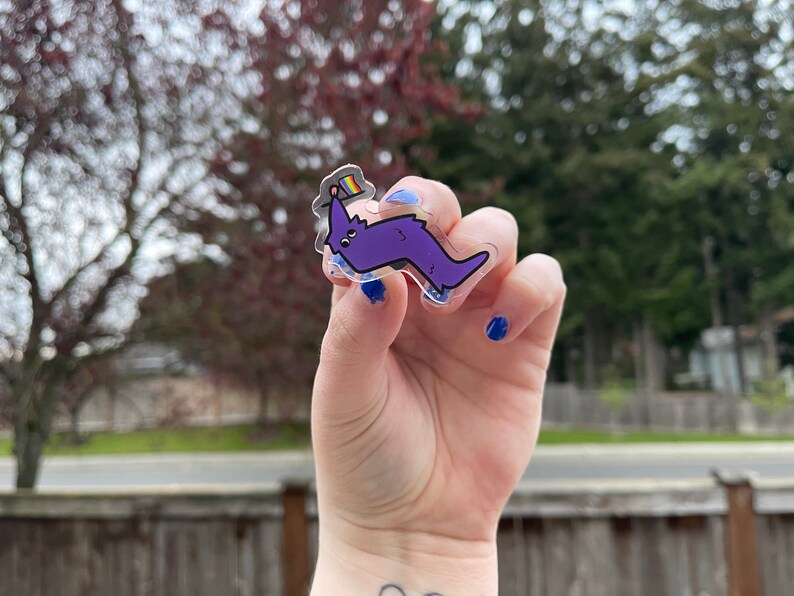 May include: A clear acrylic sticker with a purple cartoon worm wearing a rainbow flag hat. The sticker is being held by a hand with blue nail polish.