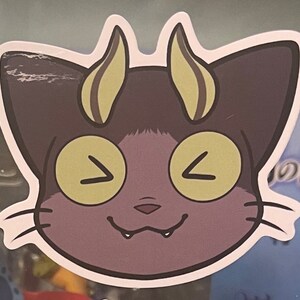 May include: A purple cat with yellow eyes and horns. The cat is smiling with its mouth open and teeth showing. The cat is a sticker.