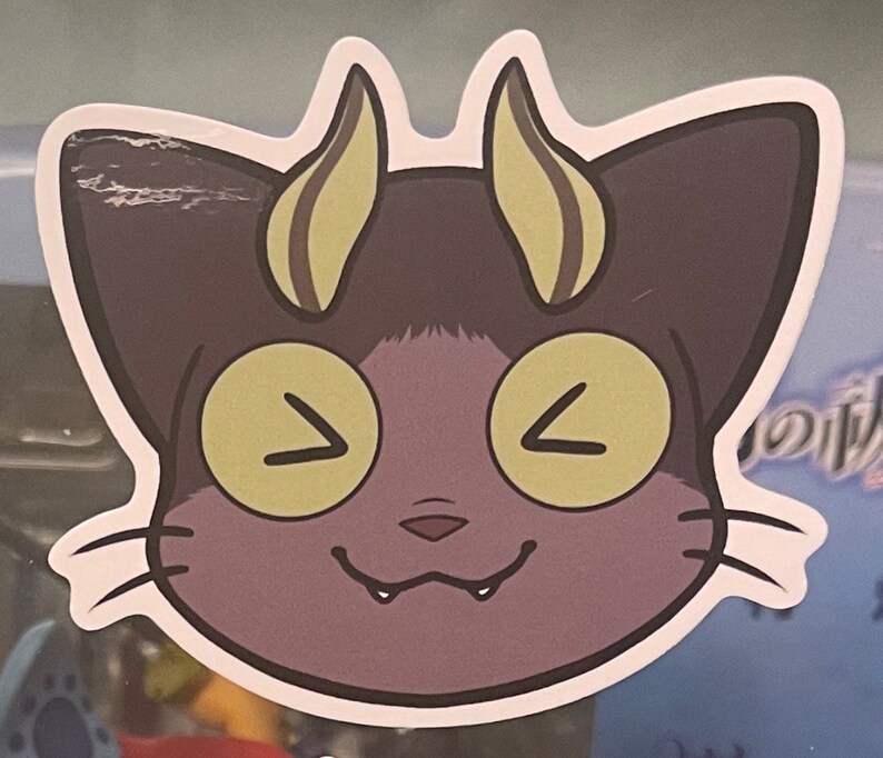 May include: A purple cat with yellow eyes and horns. The cat is smiling with its mouth open and teeth showing. The cat is a sticker.