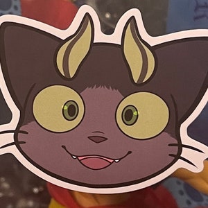 May include: A sticker of a purple cat with yellow horns and large green eyes. The cat has a mischievous grin and sharp teeth.