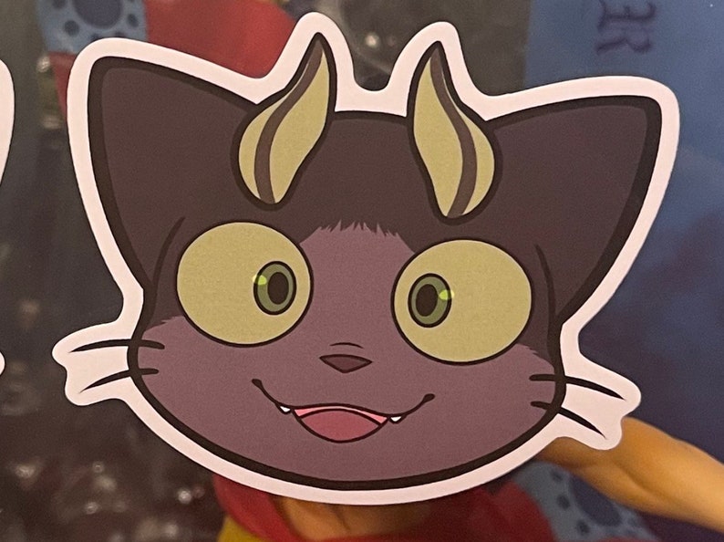 May include: A sticker of a purple cat with yellow horns and large green eyes. The cat has a mischievous grin and sharp teeth.