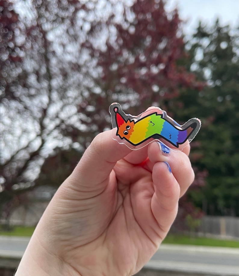 May include: A rainbow-colored worm sticker with a happy face and a party hat. The sticker is made of clear vinyl and has a glossy finish.