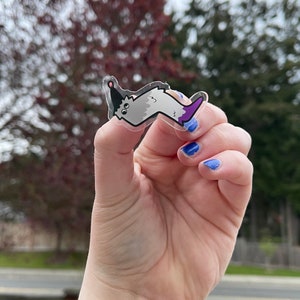 May include: A clear sticker with a cartoon worm wearing a party hat. The worm is gray with a purple tail. The sticker is being held by a hand with blue nail polish.