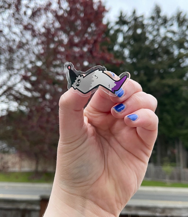 May include: A clear sticker with a cartoon worm wearing a party hat. The worm is gray with a purple tail. The sticker is being held by a hand with blue nail polish.