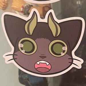 May include: A cartoon sticker of a purple cat with large green eyes and a surprised expression. The cat has two yellow horns and white whiskers.