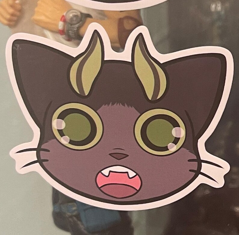 May include: A cartoon sticker of a purple cat with large green eyes and a surprised expression. The cat has two yellow horns and white whiskers.