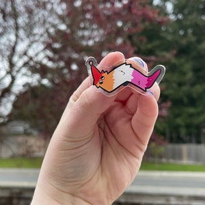 May include: A clear sticker of a cartoon worm with a red, orange, white, and pink color scheme. The worm is smiling and has a small red hat on its head.