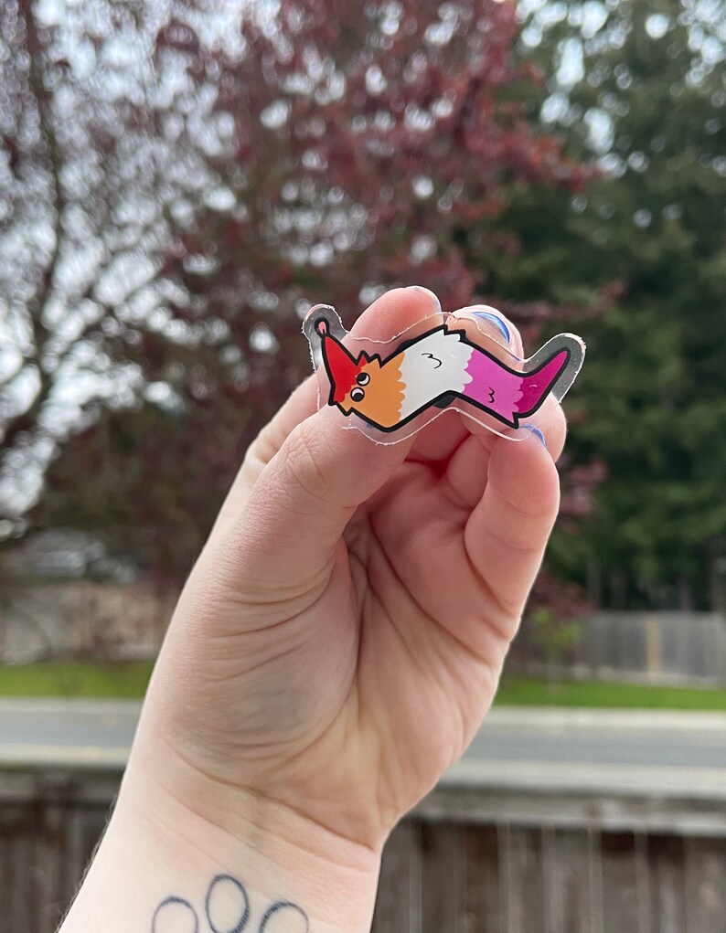 May include: A clear sticker of a cartoon worm with a red, orange, white, and pink color scheme. The worm is smiling and has a small red hat on its head.