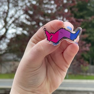 May include: A clear plastic charm shaped like a worm with a pink and purple gradient. The worm has a face with two large eyes and a small mouth. The charm is attached to a silver ring.