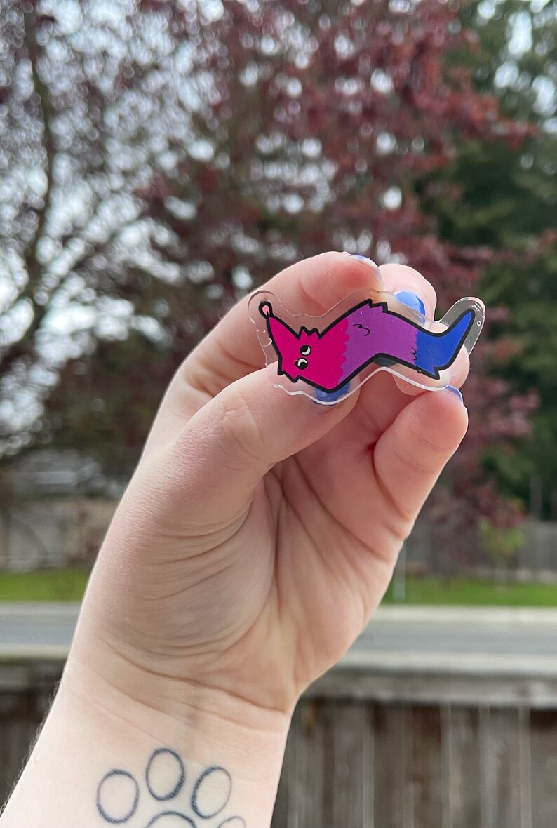 May include: A clear plastic charm shaped like a worm with a pink and purple gradient. The worm has a face with two large eyes and a small mouth. The charm is attached to a silver ring.