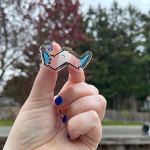 May include: A clear sticker with a cartoon worm-like creature. The creature is pink and blue with a white belly and a blue tail. The sticker is being held by a hand.