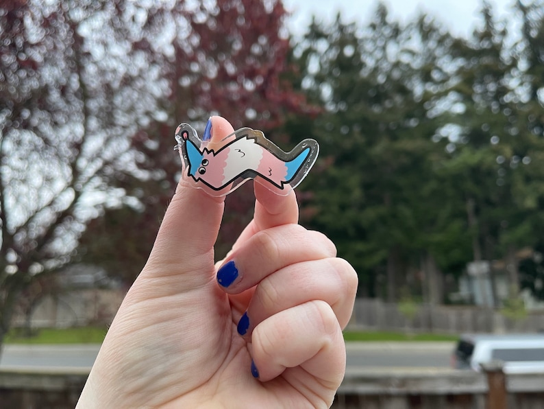 May include: A clear sticker with a cartoon worm-like creature. The creature is pink and blue with a white belly and a blue tail. The sticker is being held by a hand.