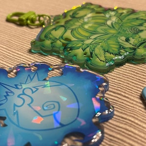 May include: Two iridescent acrylic keychains. One is blue with a white design, the other is green with a floral pattern. Both have matching colored clasps. The keychains are on a textured, neutral background.