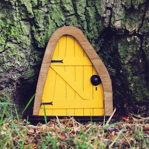 May include: A bright yellow fairy door with a wooden frame and black hinges and handle is set against a textured tree trunk. The door has a keyhole and is surrounded by grass. The words "Fairytale Doors" are in the bottom right corner.