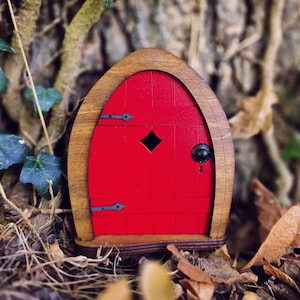 Puede incluir: Una pequeña puerta de hadas roja con marco de madera y herrajes negros. La puerta tiene una mirilla en forma de diamante y un pequeño pomo negro. El texto "Fairytale Doors" está en la parte inferior de la imagen.