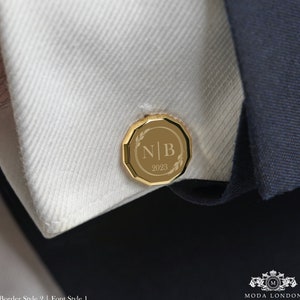 May include: Gold cufflinks with a circular design featuring the initials "N|B" and the year "2023" engraved inside a laurel wreath. The cufflinks have a faceted border.