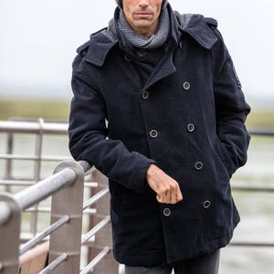 May include: A man wearing a dark blue peacoat with a hood, a gray beanie, and a gray scarf. He is standing by a metal railing.