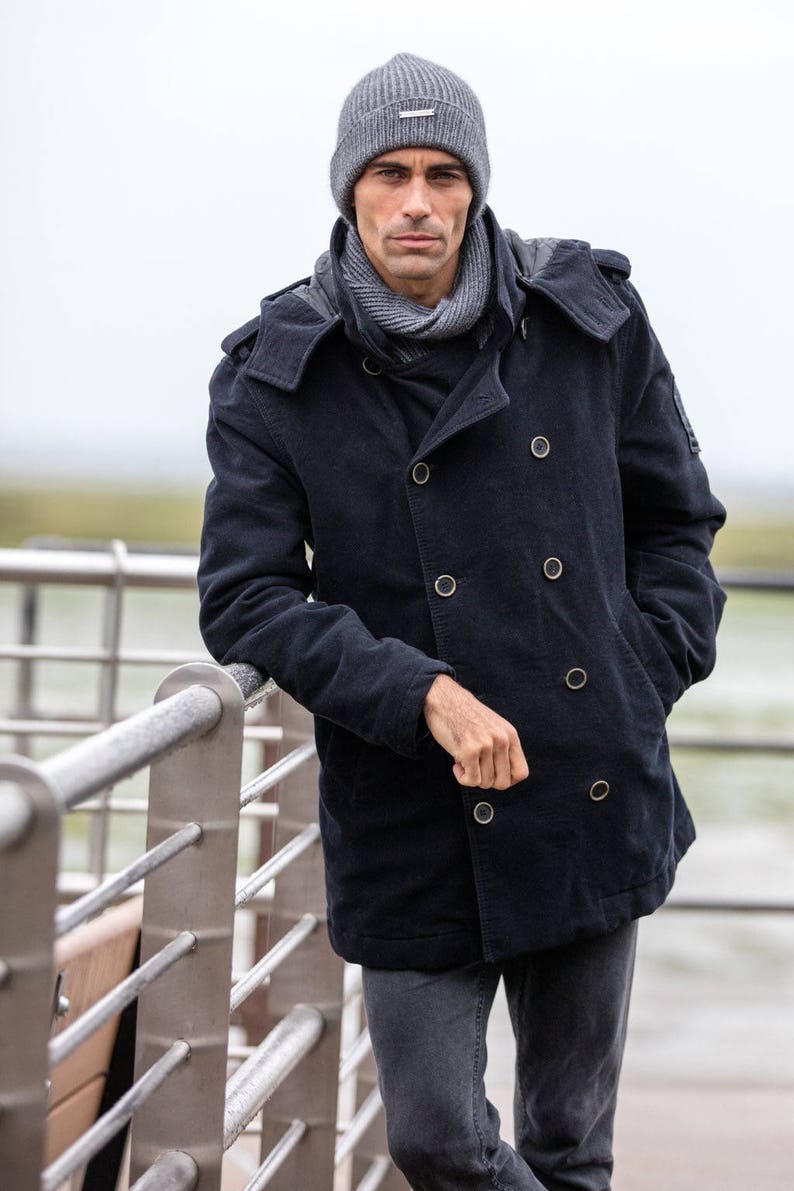 May include: A man wearing a dark blue peacoat with a hood, a gray beanie, and a gray scarf. He is standing by a metal railing.