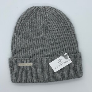 May include: Gray knitted beanie with a ribbed texture. The beanie has a small metal tag with the text "Victoria Chester" on the front.
