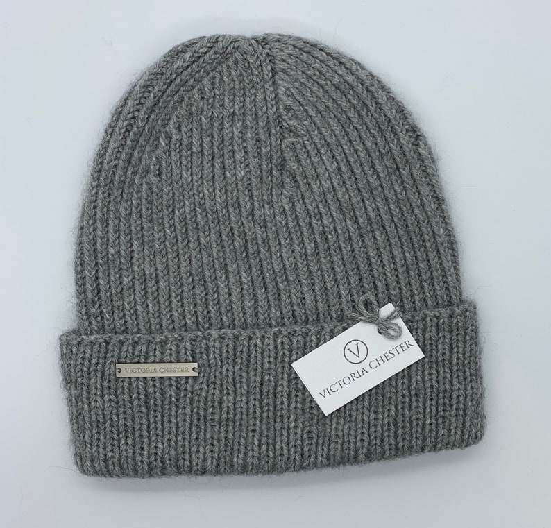May include: Gray knitted beanie with a ribbed texture. The beanie has a small metal tag with the text "Victoria Chester" on the front.