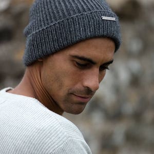May include: A man wearing a gray knitted beanie with a silver tag that reads "Woolrich John Rich & Bros." The beanie has a ribbed texture.