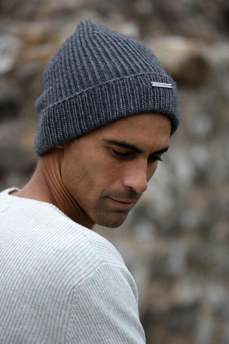 May include: A man wearing a gray knitted beanie with a silver tag that reads "Woolrich John Rich & Bros." The beanie has a ribbed texture.