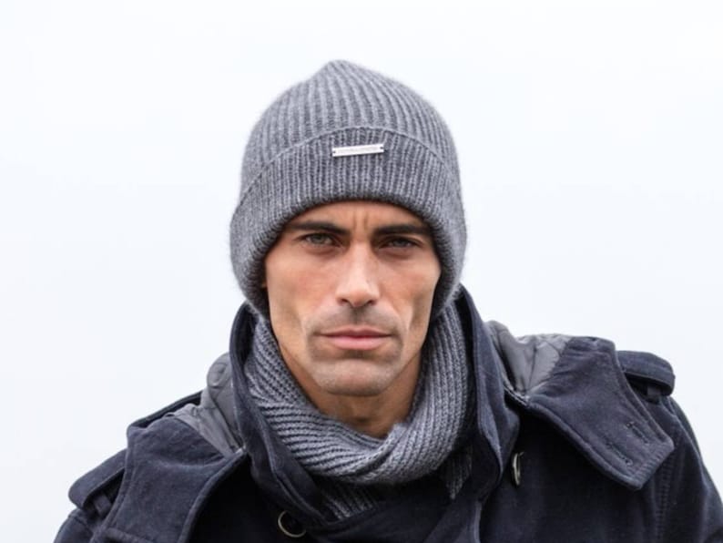 May include: A gray knitted beanie with a silver logo on the front. The beanie is worn by a person with a gray scarf and a dark blue jacket.