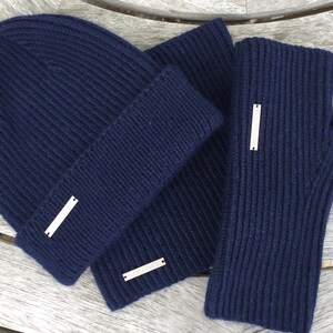 May include: A navy blue knit beanie, scarf, and fingerless gloves set. The set features a silver metal tag on each piece.