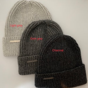 May include: Three knitted beanies in different shades of gray: light gray, dark gray, and charcoal. Each beanie has a small label with the text "Victoria Chester" on it.