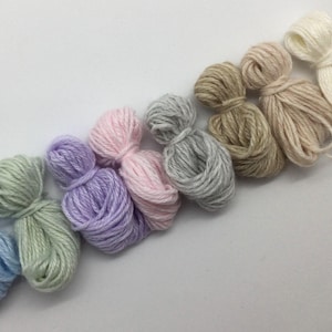 Op de afbeelding: Een verzameling kleurrijke garenbundels in tinten blauw, groen, paars, roze, grijs, beige en wit. Het garen is diagonaal geplaatst tegen een witte achtergrond, met een verscheidenheid aan zachte pastelkleuren. Ideaal voor knutsel- en breiprojecten.