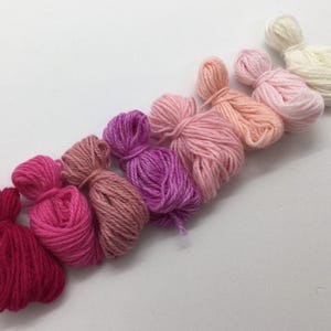 Puede incluir: Una colección de ocho ovillos de hilo en tonos de rosa, rojo y blanco. El hilo está dispuesto en un degradado de rojo oscuro a blanco.